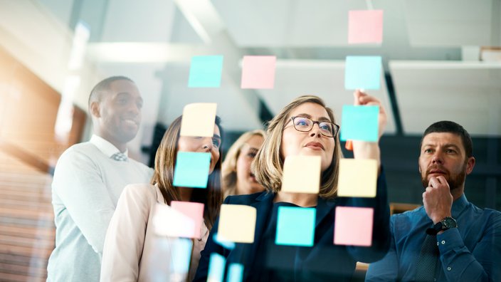 Brainstorming mit Post-its Das Bild zeigt eine Gruppe von Menschen vor einer Glaswand, die mit bunten Haftnotizen bedeckt ist. Die Personen scheinen in einer Besprechung oder Brainstorming-Sitzung zu sein und schauen auf die Notizen, während sie diskutieren und Ideen austauschen. Die Haftnotizen sind in verschiedenen Farben wie Blau, Gelb und Rosa. Die Szene findet in einem modernen Büro oder Konferenzraum mit heller Beleuchtung statt.